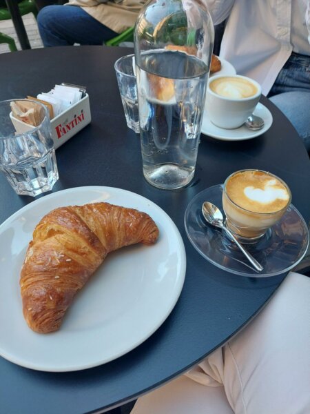 Ein Croissant mit einem Espresso Macchiato, einer Wasserkaraffe und im Hintergrund noch ein Croissant und eine Tasse Cappuccino