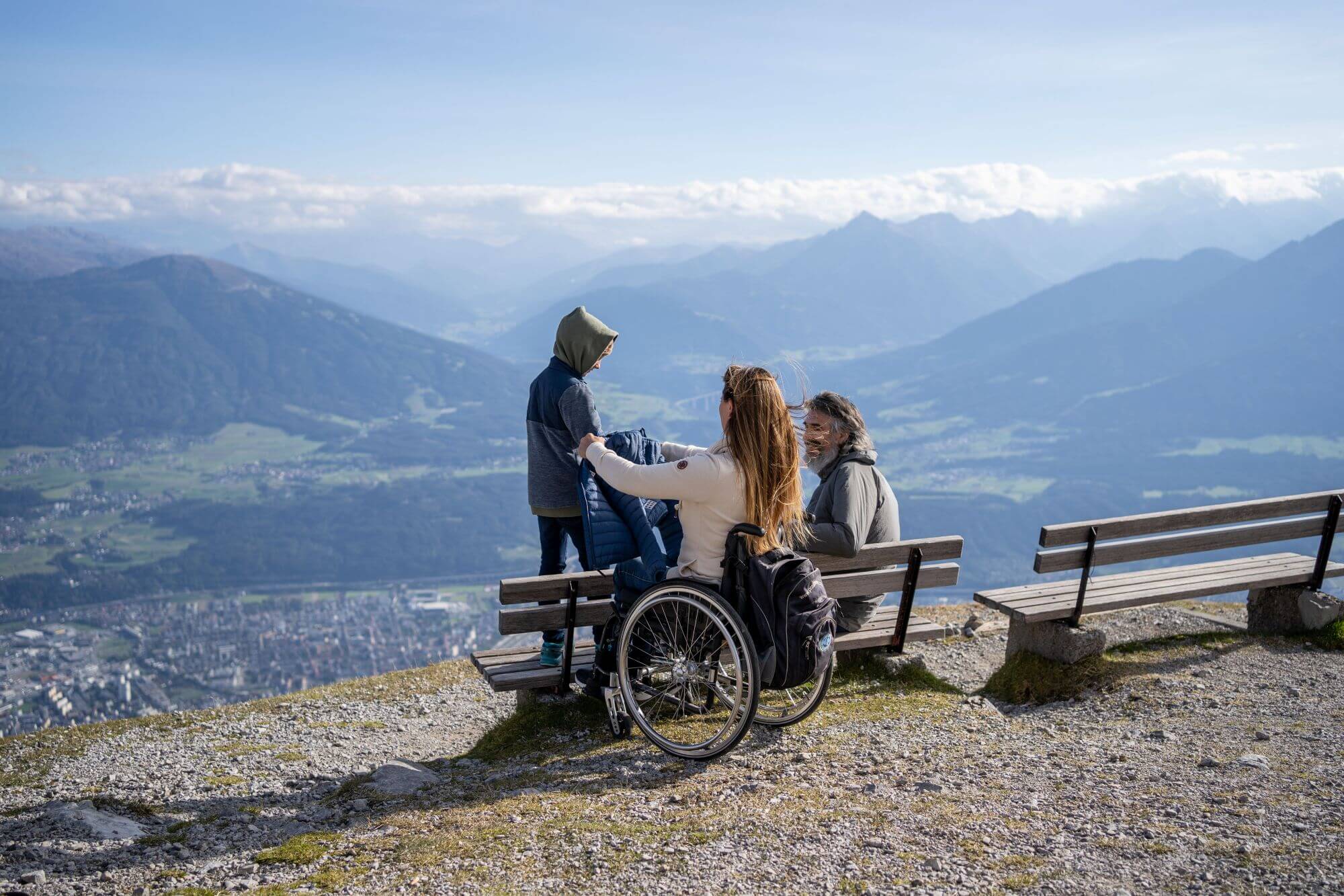 Eine Frau im Rollstuhl, ein Kind und ein Mann befinden sich um eine Sitzbank am Hafelekar in Innsbruck, dahinter sieht man ins Tal und über Berge
