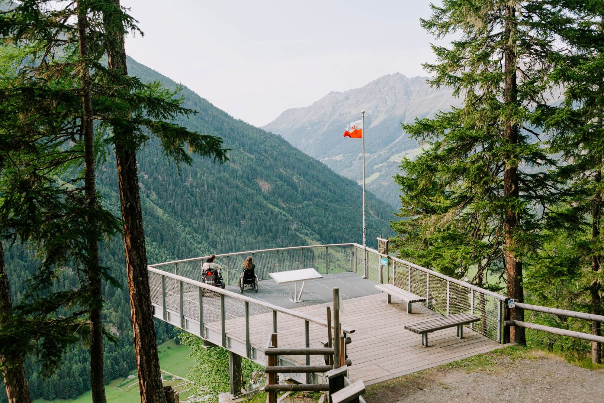 Zwei Menschen im Rollstuhl auf einer Aussichtsplattform, die Tiroler Flagge weht auf einem Mast, dahinter grüne Berge