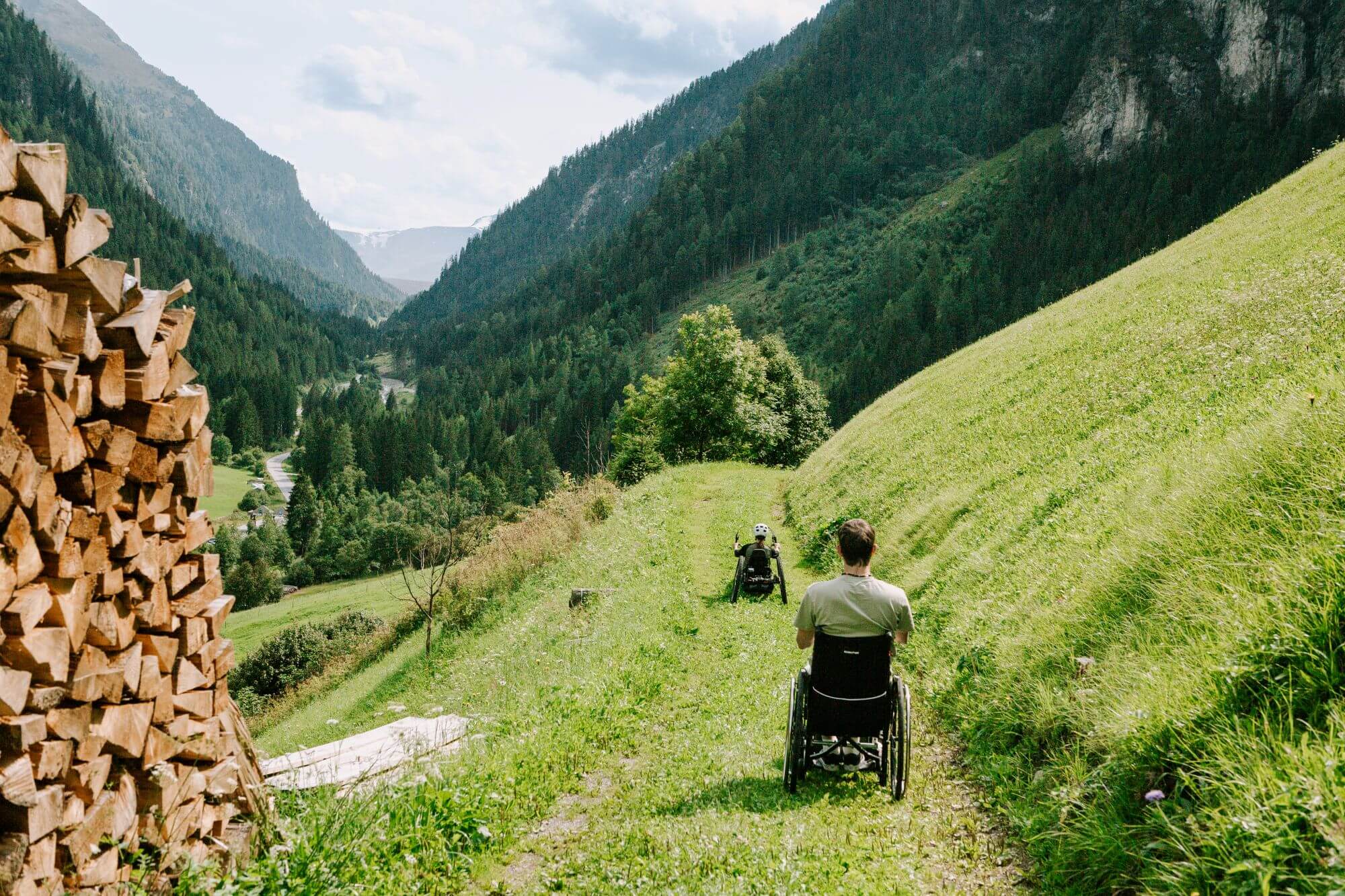Zwei Menschen im Rollstuhl auf einem Wanderweg in einer Wiese, links ein Stapel Holz, im Hintergrund grüne Berge