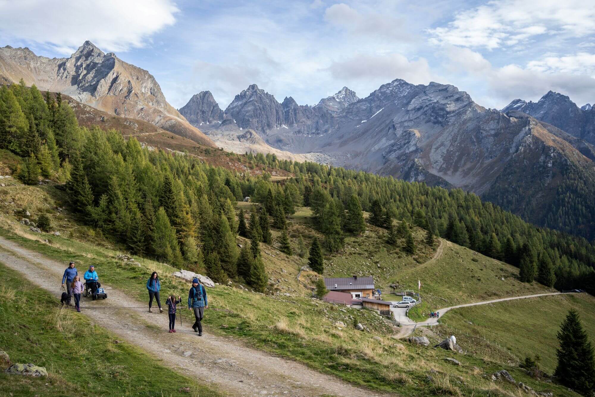 Eine Gruppe auf einem Wanderweg, einer sitzt in einem Rollstuhl. Dahinter ist eine Hütte, Wald, grüne Wiese und berge