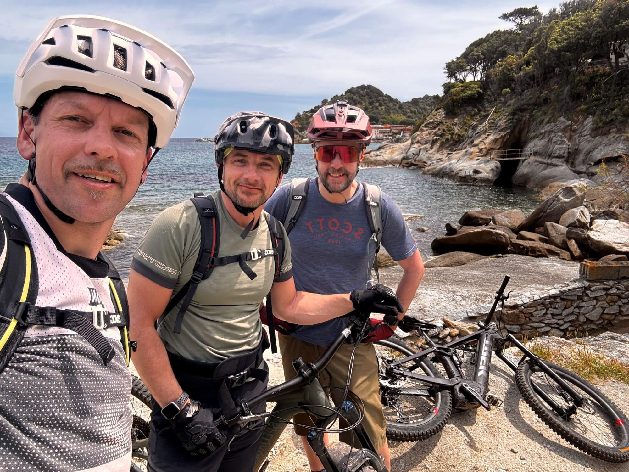 Selfie von drei Mountainbikern auf Elba vor dem Meer, daneben liegt ein Mountainbike
