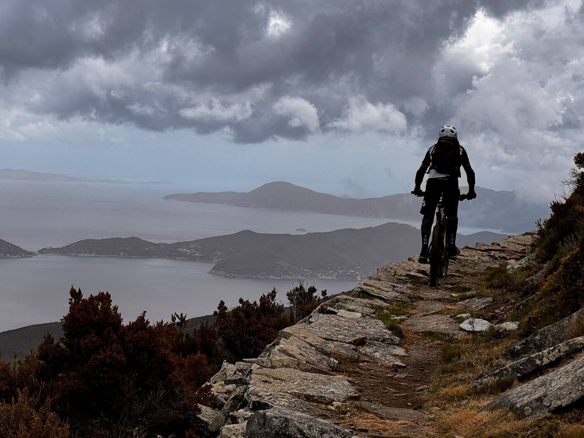 Mountainbiker auf Elba fährt auf schmalem Steinweg, im Hintergrund sieht man das Meer, es ist bewölkt.