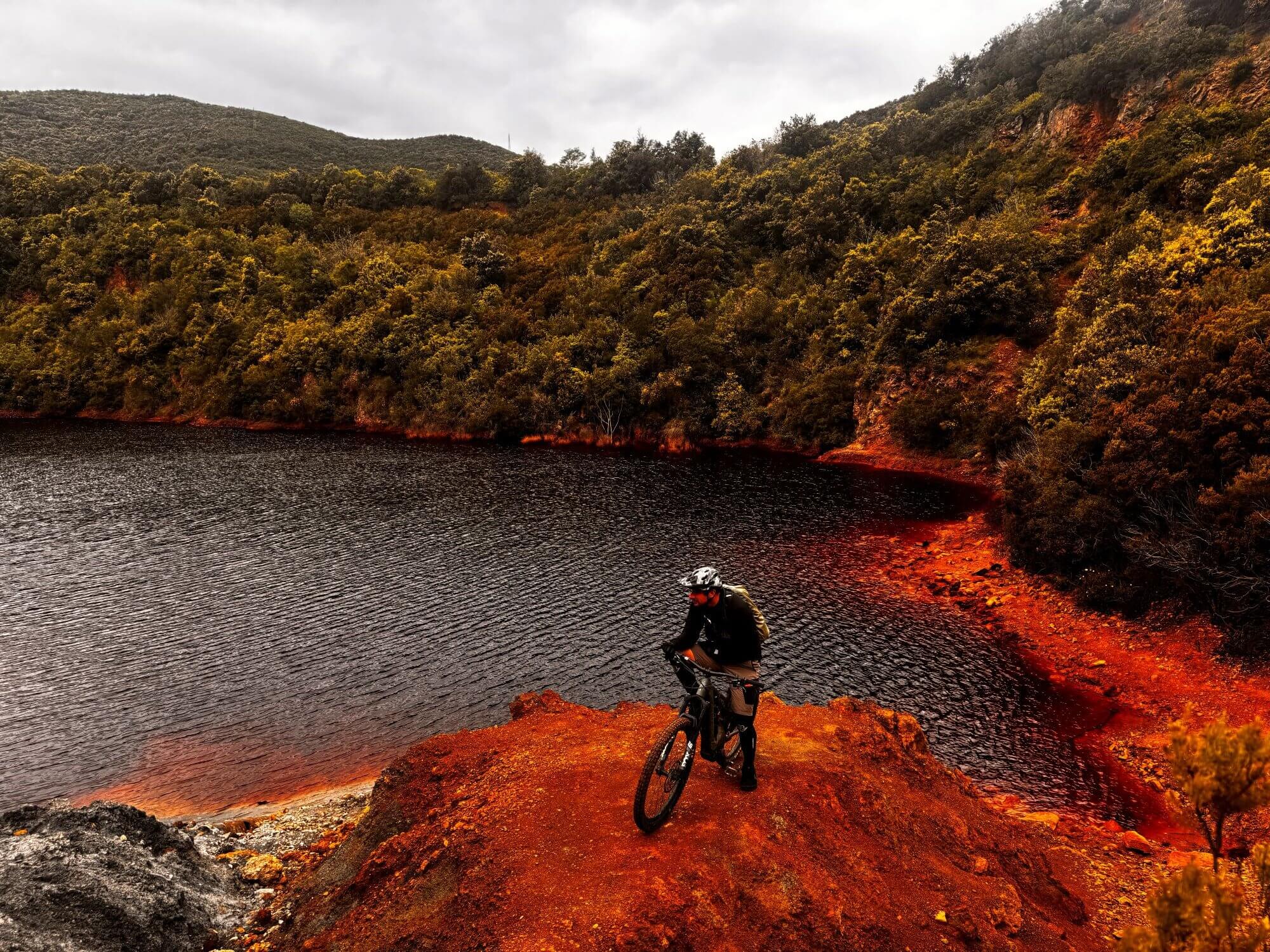Mountainbiker steht auf roter Klippe. dahinter ein dunlkler See auf Elba