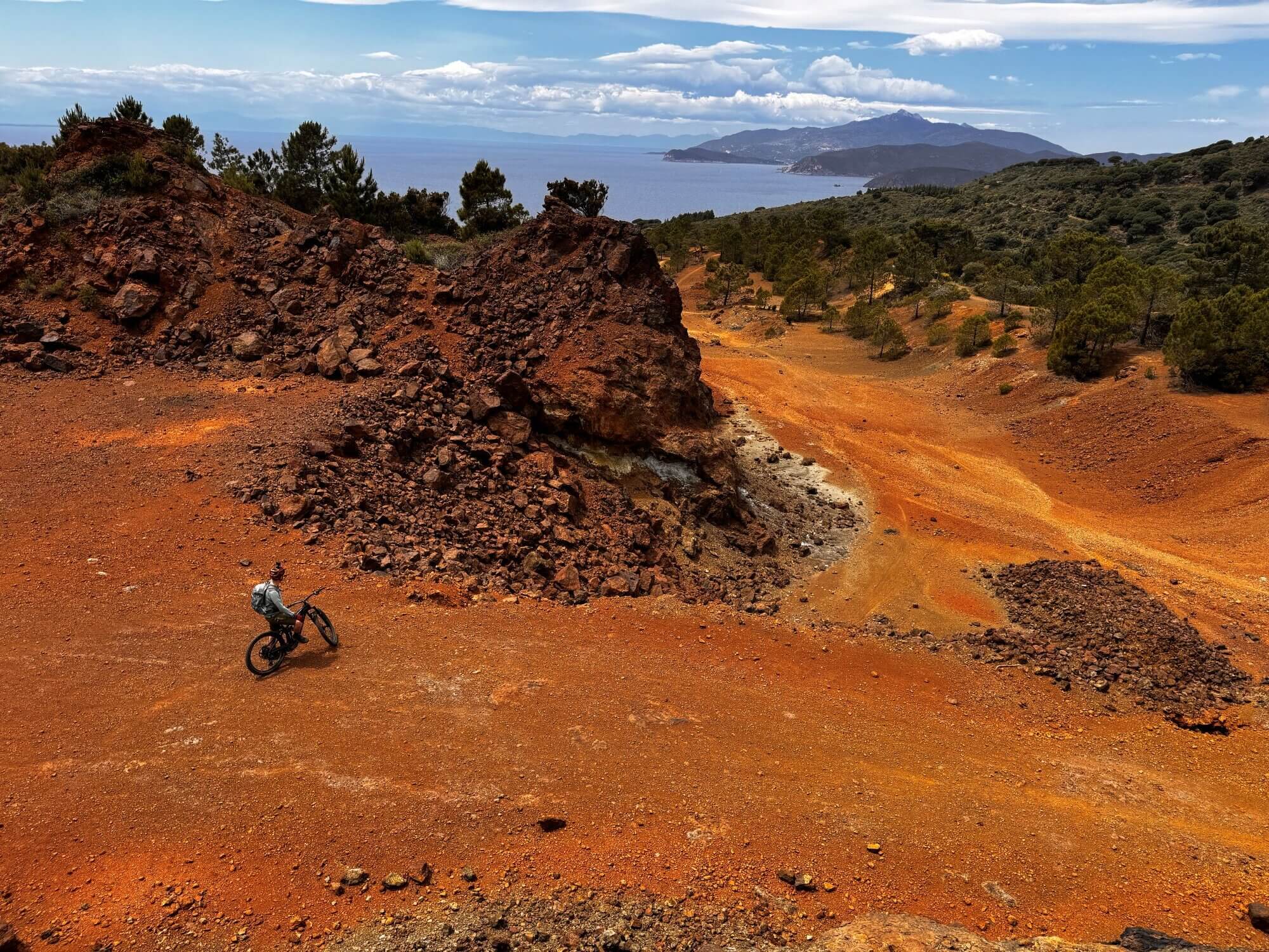 Mountainbiker auf Elba fährt auf roter Erde, dahinter das blaue Meer