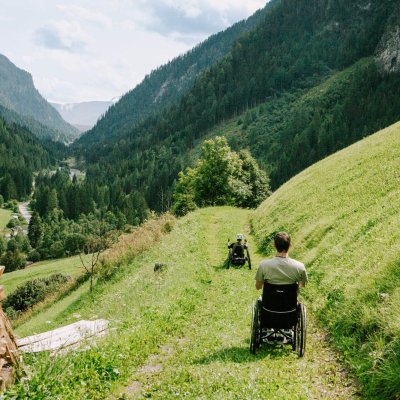 Zwei Menschen im Rollstuhl auf einem Wanderweg in einer Wiese, links ein Stapel Holz, im Hintergrund grüne Berge